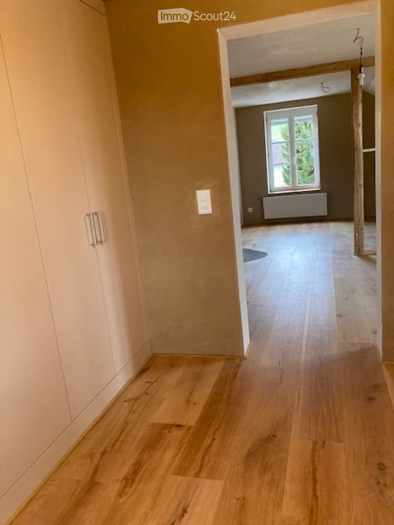 Empty hallway with light wooden flooring, beige walls, and a closed white cabinet on the left. The hallway leads to another room with a window and radiator.