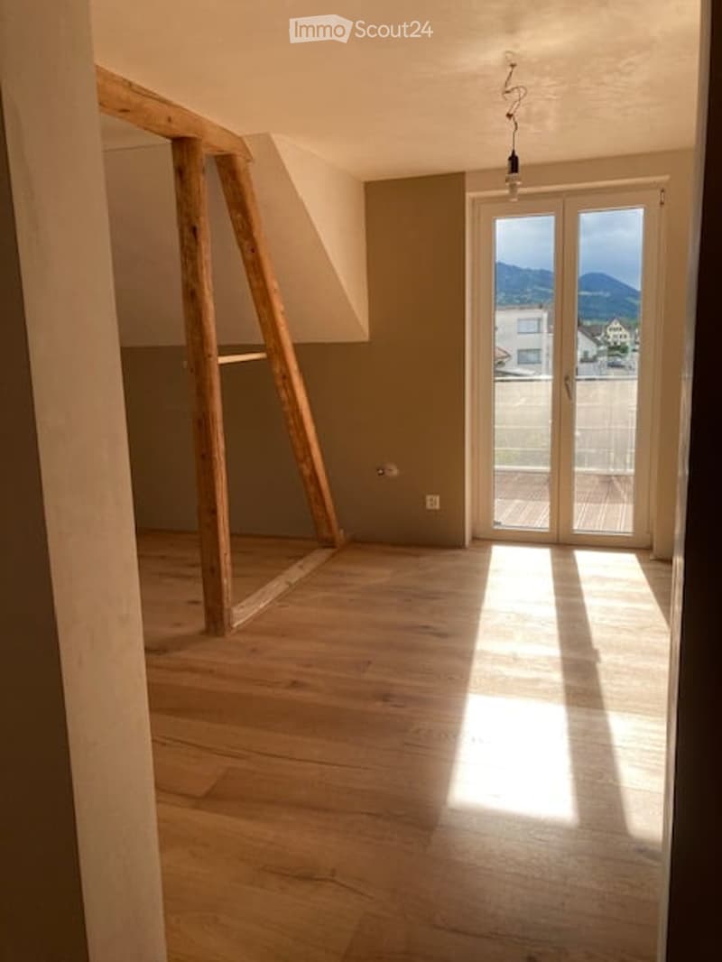 Empty room with wooden flooring, exposed wooden beams, large glass door leading to a balcony with a scenic mountain view.