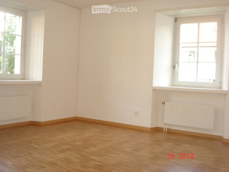 Empty room with wooden flooring, white walls, and two windows with white frames. Radiators are present on both walls.