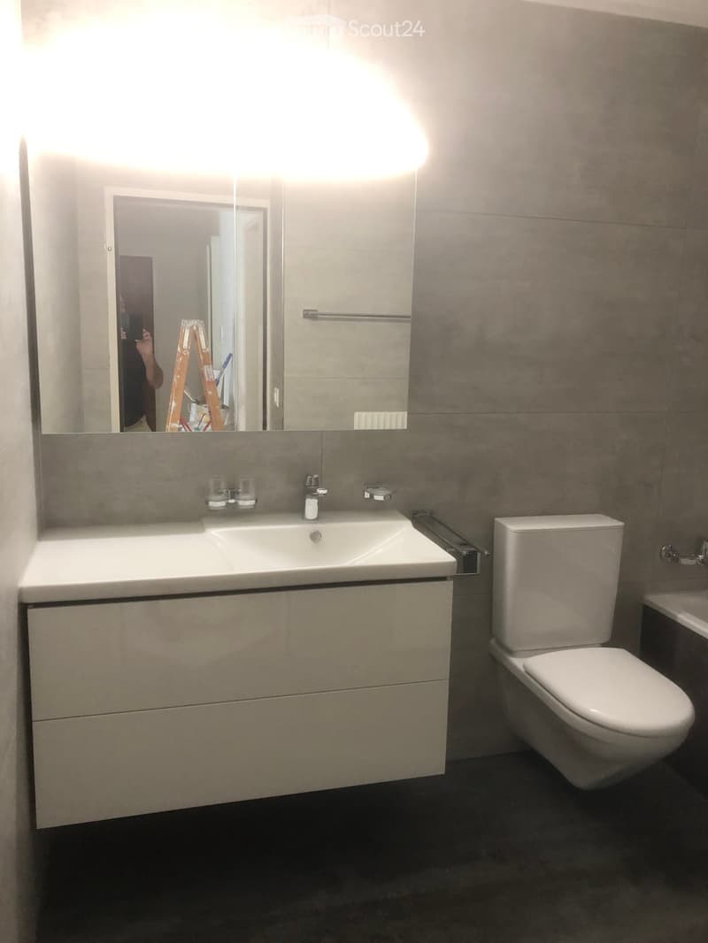 Modern bathroom with a white sink, toilet, and large mirror. The sink has a modern faucet and two cups on the counter. The walls are tiled in a neutral color.