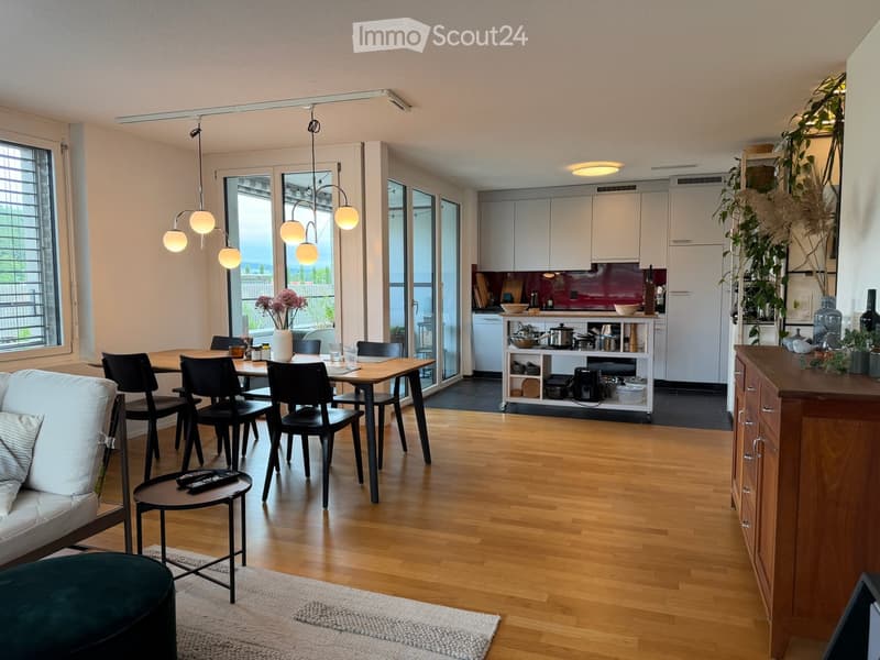 Modern open-plan living and dining area with a sofa, ottoman, coffee table, dining table with chairs, and a fully equipped kitchen with cabinets, appliances, and pendant lighting.