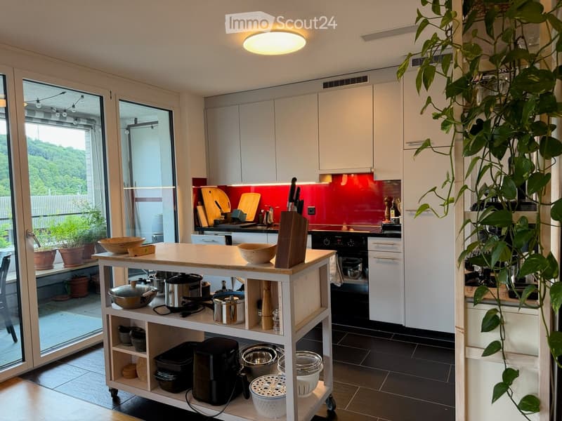 Modern kitchen with white cabinets, a red backsplash, and a central island. The island features bowls, a cutting board, and various kitchen utensils. A large plant is visible on the right side, and the kitchen has large windows with a view of greenery out
