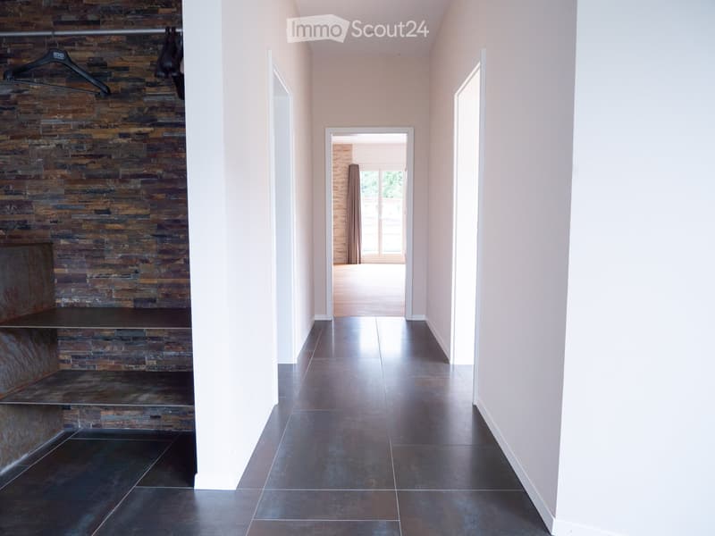 Modern hallway with dark tiled flooring, light-colored walls, and a stone accent wall on the left. A coat rack with hanging items is visible, and an open doorway leads to another room with a curtained window.