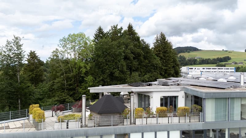 Modern building with a flat roof, solar panels, and a small gazebo surrounded by potted plants and trees, overlooking a lush green landscape.