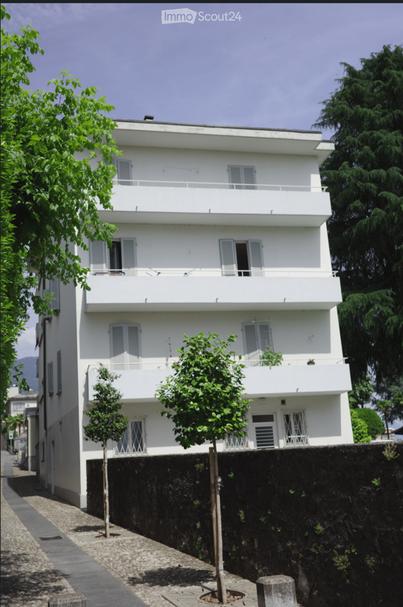 A three-story white building with multiple balconies and shutters, surrounded by trees and a stone wall.