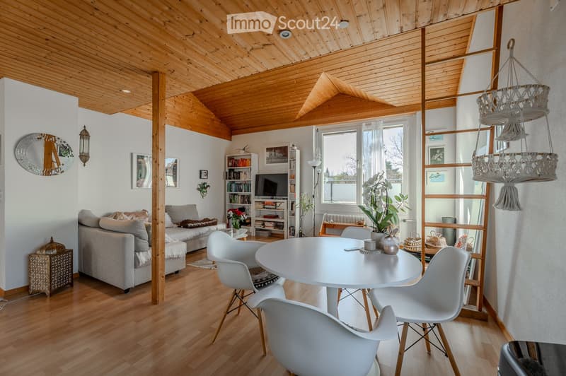 A modern living room with wooden floors, exposed wooden beams, a grey sofa, a white dining table with chairs, a bookshelf, and a television. The room features a hanging decorative chandelier and a large window with a view of the outdoors.