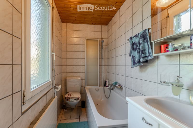 Bathroom with tiled walls and wooden ceiling, featuring a bathtub, toilet, sink, mirror, and towel hanging on the wall.