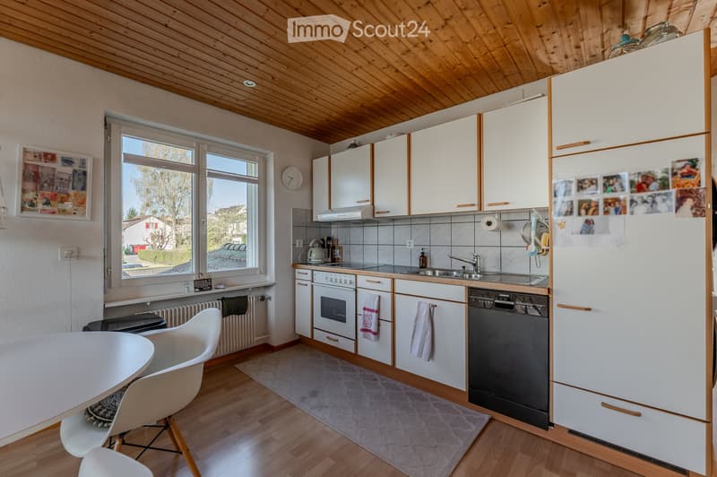 Bright kitchen with white cabinets, wooden ceiling, and large windows. Includes a refrigerator, oven, dishwasher, sink, and a small dining area with a table and chairs.