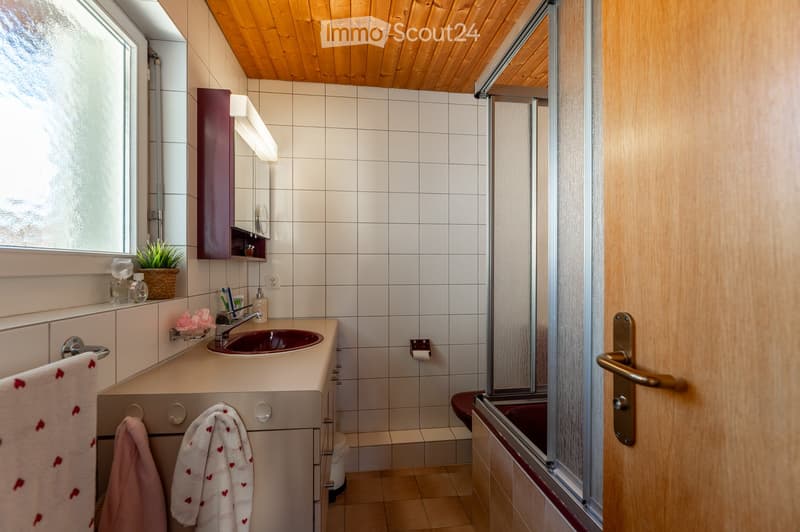 A small bathroom with white tiled walls, a wooden ceiling, a sink with a red basin, a mirror cabinet, a toilet, a shower stall, and a small plant on the windowsill.