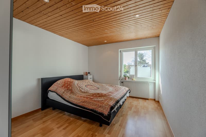 Bright bedroom with wooden flooring, a large window, a bed with a patterned bedspread, a nightstand, and a radiator beneath the window.