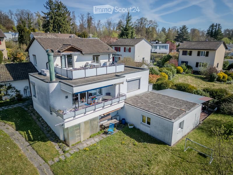 Aerial view of a modern two-story house with a flat roof, surrounded by a well-maintained garden and neighboring houses. Features include a balcony, patio area, and outdoor furniture.