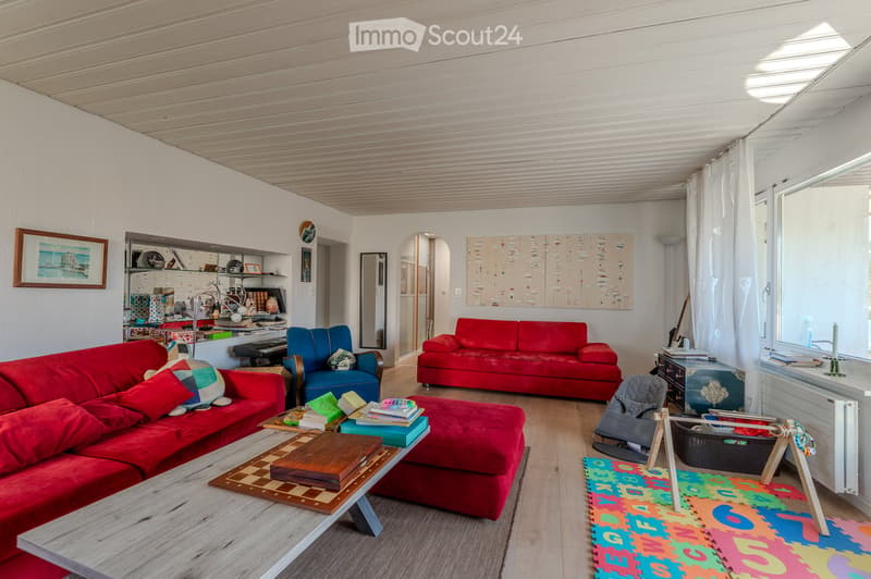 Bright living room with red sofas, a coffee table, ottoman, armchair, baby chair, and colorful play mat. Shelves and artwork are visible on the walls.
