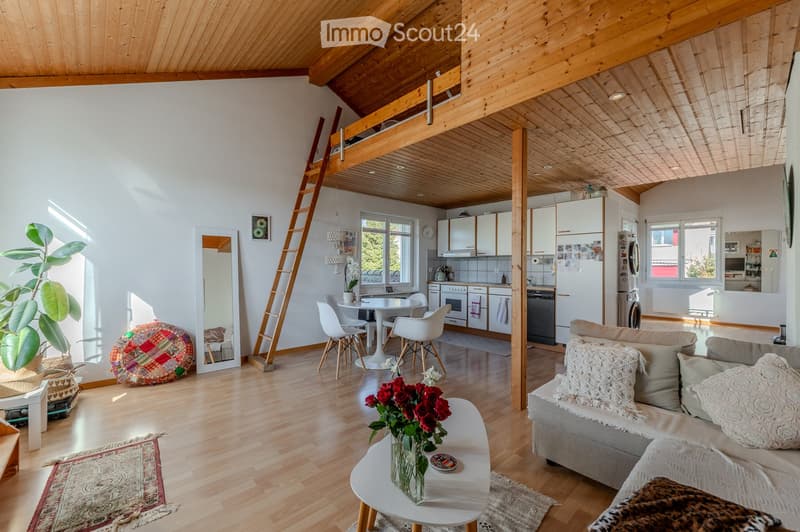 Bright, open-concept living and dining area with wooden floors, white walls, and a slanted wooden ceiling. Features a grey sofa with decorative pillows, a white coffee table with a flower vase, a dining table with white chairs, a ladder leading to a loft