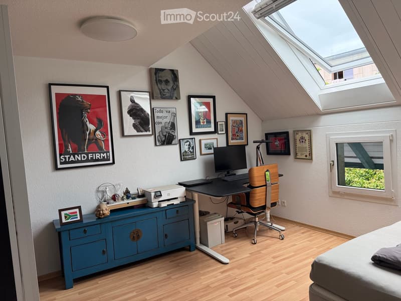 Cozy attic room with wooden flooring, slanted ceilings, and skylights. Features a desk with a chair, a printer, a blue cabinet, and a bed with pillows.