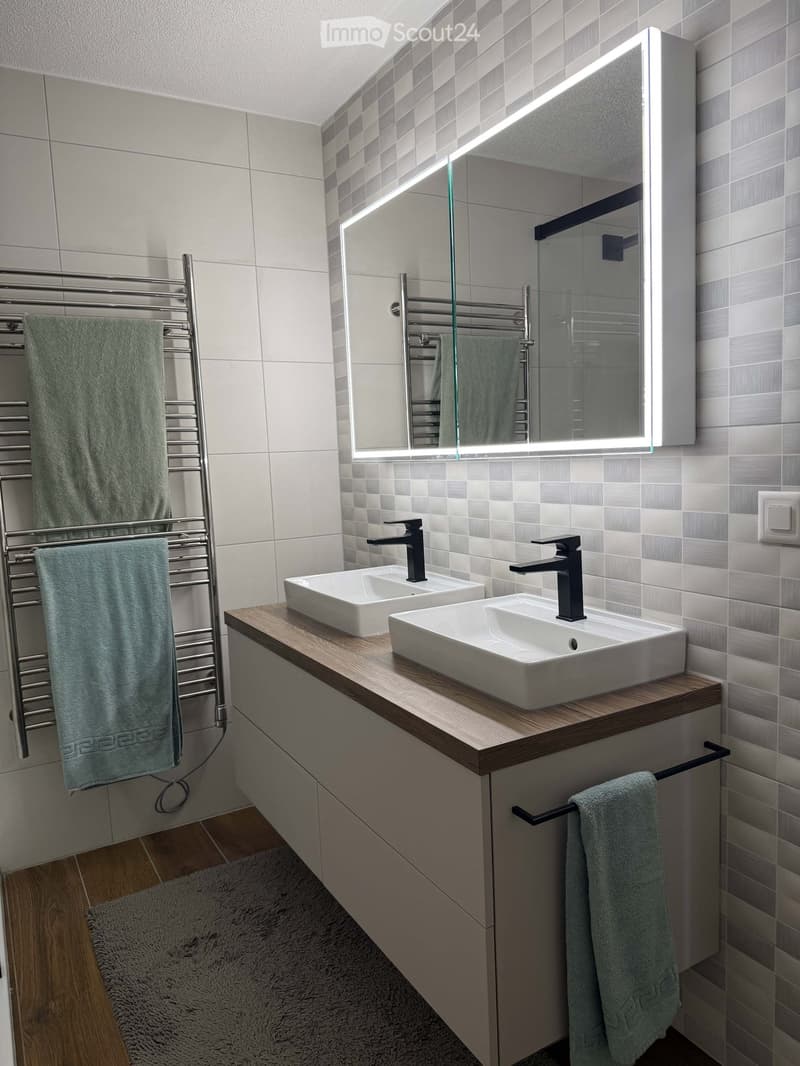Modern bathroom with two white sinks, sleek black faucets, a large illuminated mirror, a towel rack with green towels, and a wooden countertop.