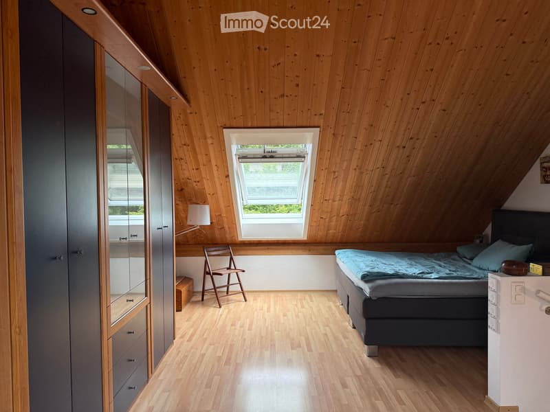 Cozy attic bedroom with wooden walls and ceiling, featuring a bed, a chair, a wardrobe, and a lamp.