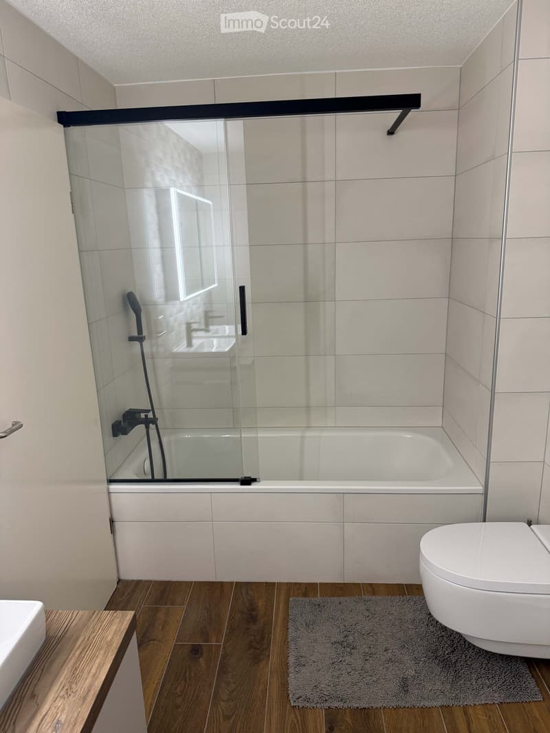 Modern bathroom with a bathtub, toilet, shower, and a wooden floor. The shower has a glass enclosure, and there is a small bathroom rug in front of the toilet.