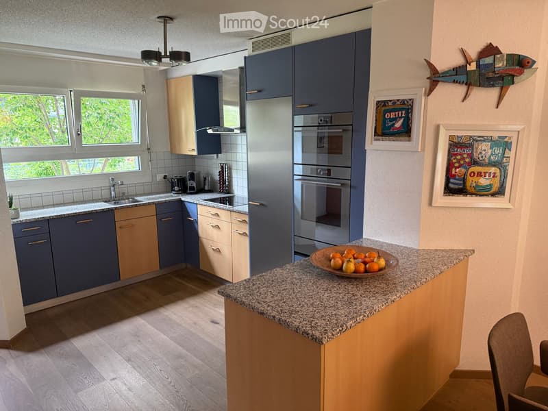 Modern kitchen with blue and beige cabinets, granite countertop, stainless steel appliances, and a bowl of fruit on the island.