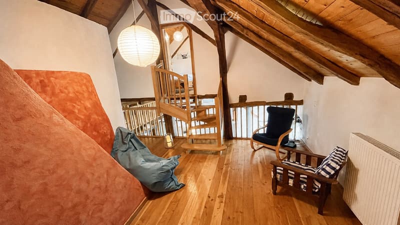 Cozy attic room with wooden floors, exposed wooden beams, a staircase leading to another level, a blue bean bag chair, a wooden armchair, a small table, and a hanging spherical lamp.