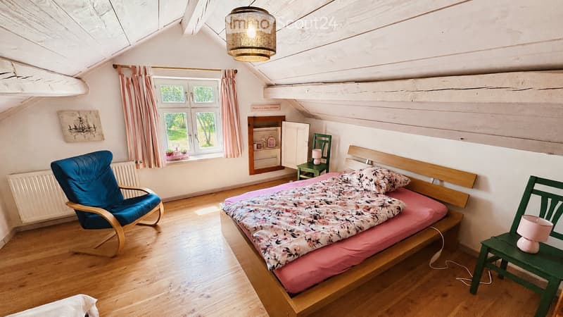 Cozy attic bedroom with wooden floors, slanted ceilings, and large windows. Features a double bed with floral bedding, a blue armchair, a small green table with a lamp, and a built-in cabinet.