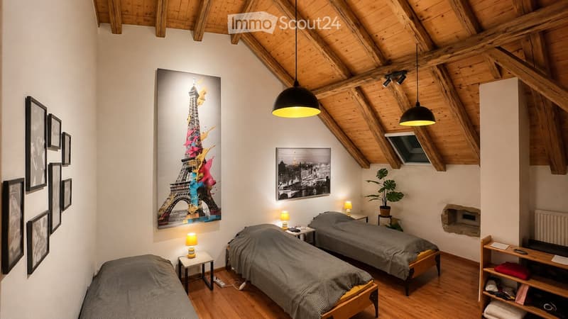 Cozy attic bedroom with wooden beams, three single beds, nightstands, table lamps, artwork, and a potted plant.