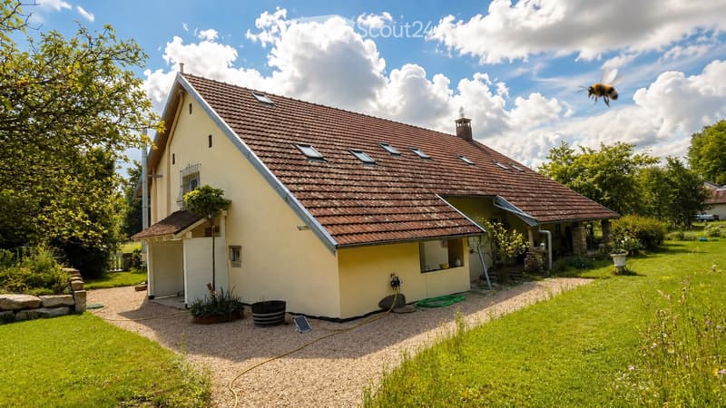 Single-story house with a brown tiled roof, yellow walls, and a small balcony. The house is surrounded by a well-maintained garden with green grass, trees, and a gravel driveway.