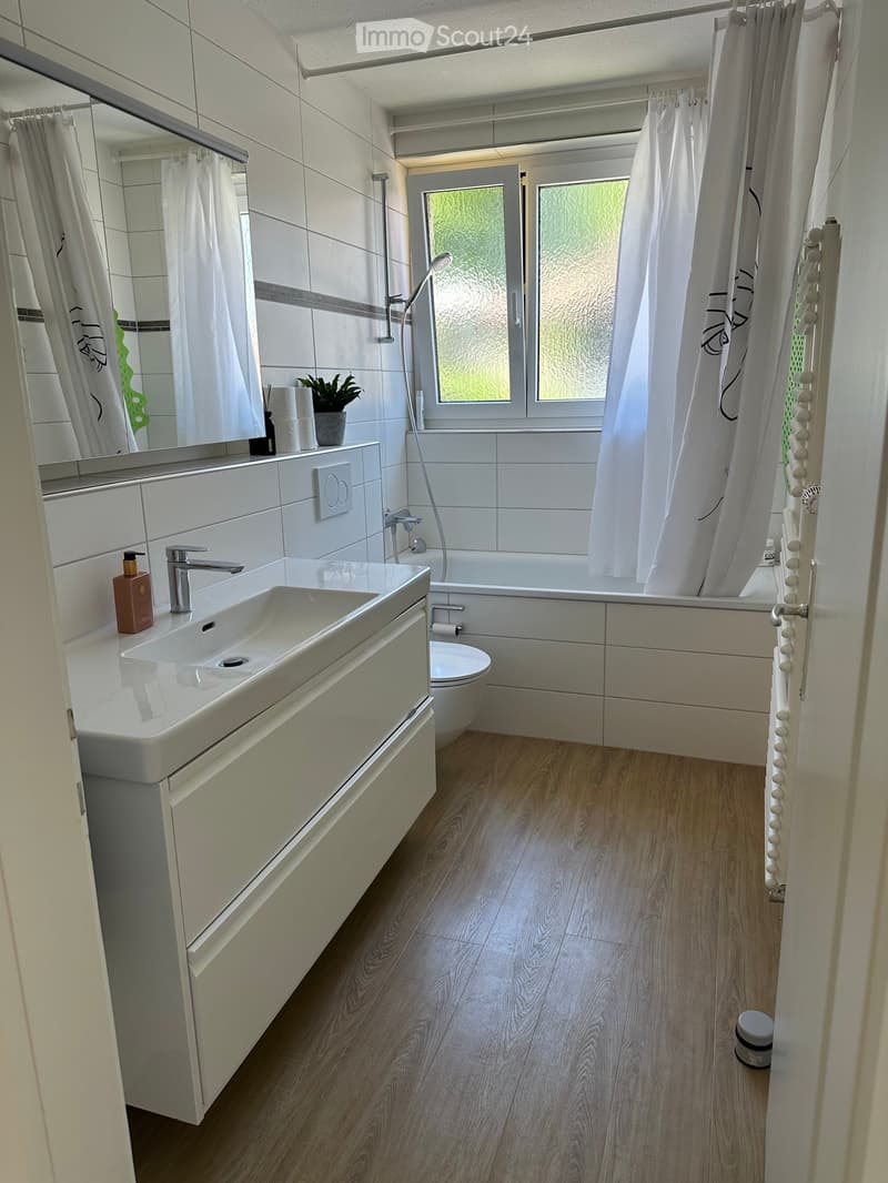 Modern bathroom with white tiles, wooden flooring, a large sink with a modern faucet, a bathtub with a shower curtain, a toilet, and a small plant on the windowsill.