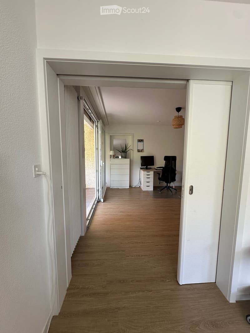A clean, modern hallway with wooden flooring, white walls, and a hanging woven pendant light. An open doorway leads to a furnished room with a desk, chair, and computer.