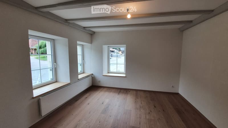 Empty room with wooden flooring, white walls, and multiple windows with sills. Exposed wooden beams on the ceiling and a single light fixture.