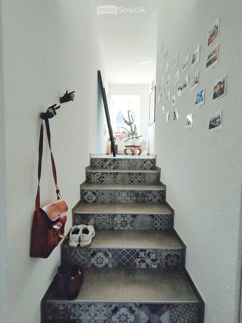 Staircase with patterned tiles, white walls, and a small plant visible at the top. Shoes and a bag are placed on the stairs.