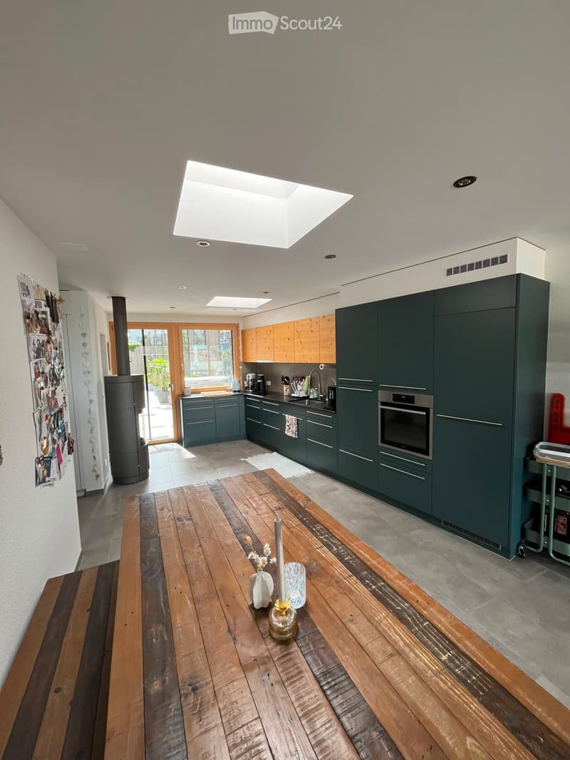Open-plan kitchen and dining area with dark green cabinets, wooden dining table, and modern appliances.