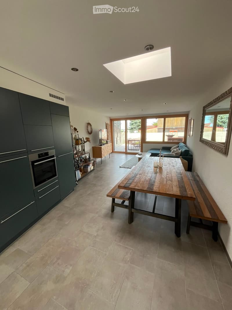 Open-plan kitchen and dining area with a large wooden dining table, benches, a sofa, dark green cabinets, and a modern oven. Large windows and a skylight provide natural light.