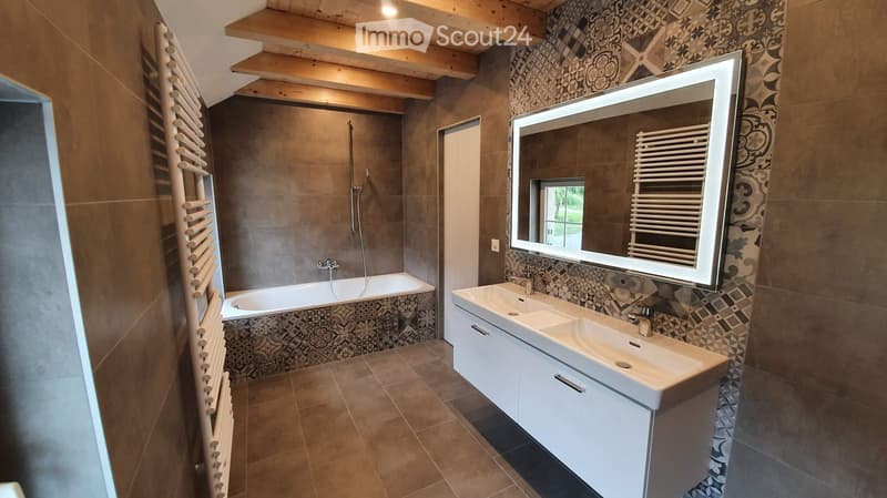 Modern bathroom with a large mirror, double sink, bathtub, and radiator. The walls are tiled with a decorative pattern, and the floor is tiled in a warm brown tone.