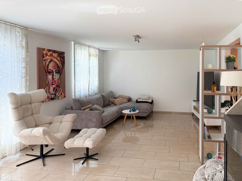 Modern living room with a grey sofa, beige armchair, small round table, wooden shelf, and large painting on the wall.