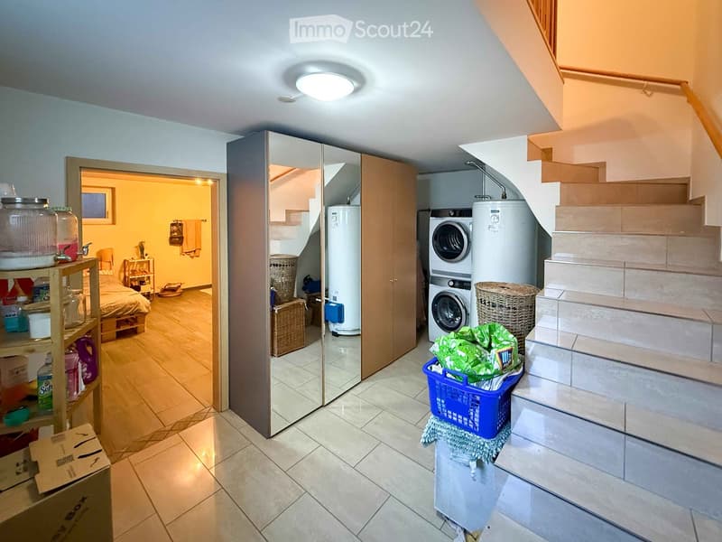 Hallway with tiled floor, staircase leading upwards, washing machine, water heater, and a partially open closet with a mirror. Laundry items and cleaning supplies are visible.