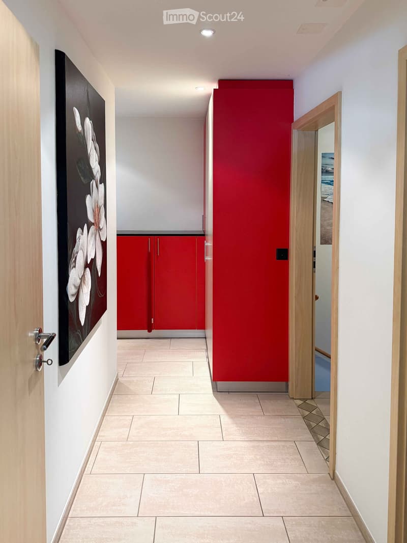 Modern hallway with beige tiled flooring, white walls, and red accent wall. Features a large framed floral artwork on the left and a partially visible red cabinet with silver handles on the right.