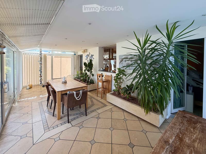 Bright, airy outdoor dining area with a large wooden table, four chairs, and two stools, surrounded by potted plants and large windows.