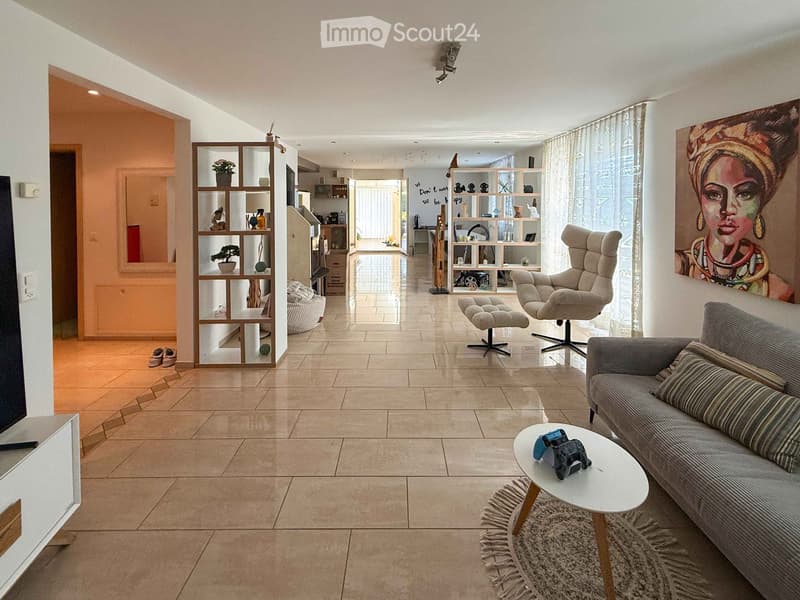 Modern living room with tiled flooring, large windows, a grey sofa, coffee table, armchair, and decorative shelves.
