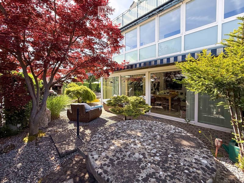 Garden area with a wicker chair, a stone table, and a small table with chairs inside the house, surrounded by trees and shrubs.