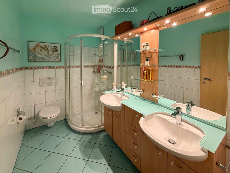 Modern bathroom with two sinks, a toilet, a glass-enclosed shower, and a wooden vanity with a mirror and shelves. The walls are painted in a light green color with decorative tiles along the lower half.