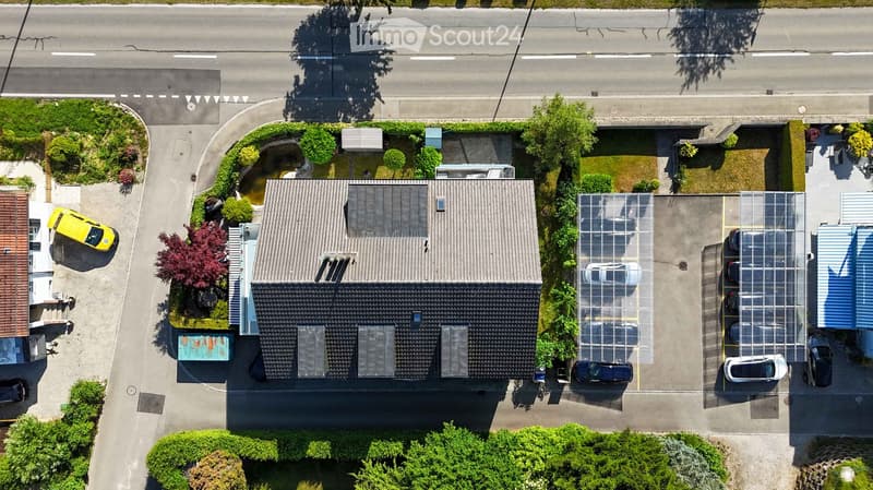 Aerial view of a residential building with a grey roof, multiple windows, and a parking area with several cars.