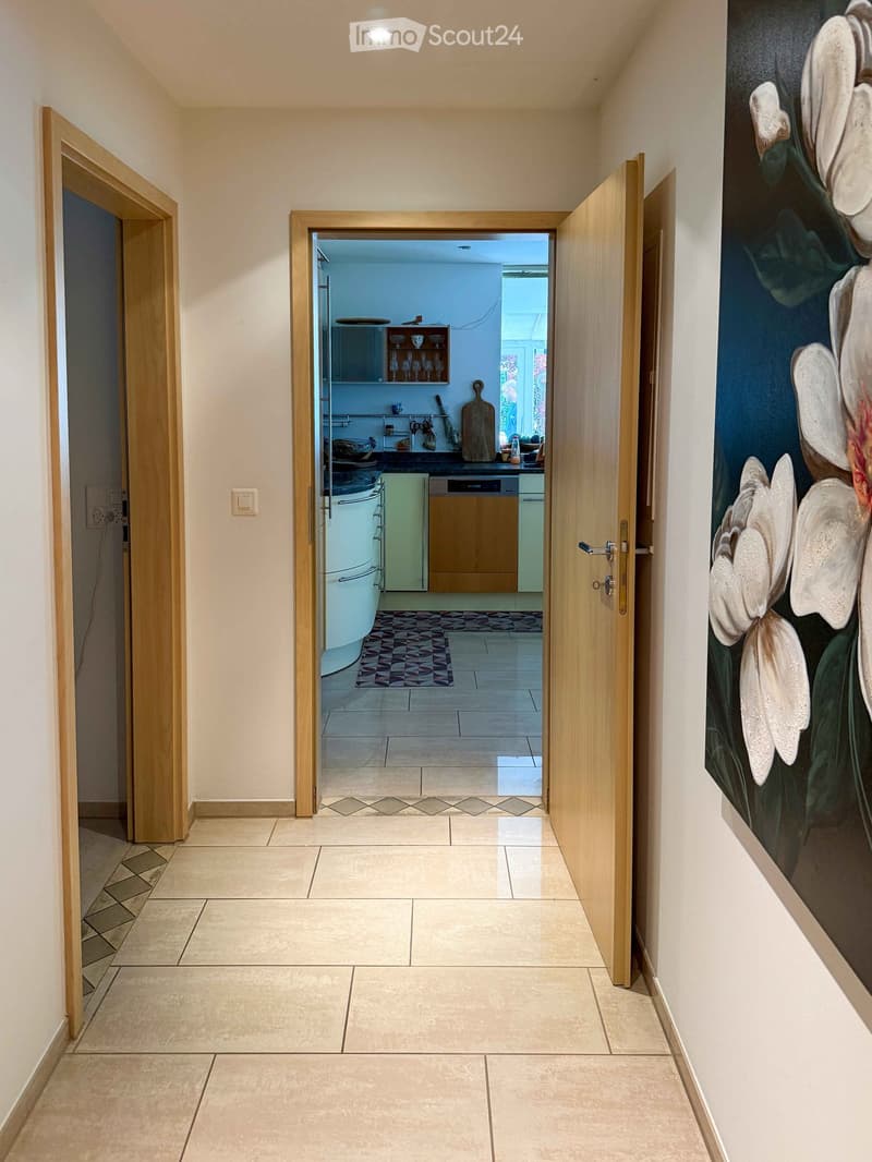 Bright hallway with tiled flooring, wooden doors, and a large floral painting on the right wall. The hallway leads to an open kitchen area visible through an open doorway.