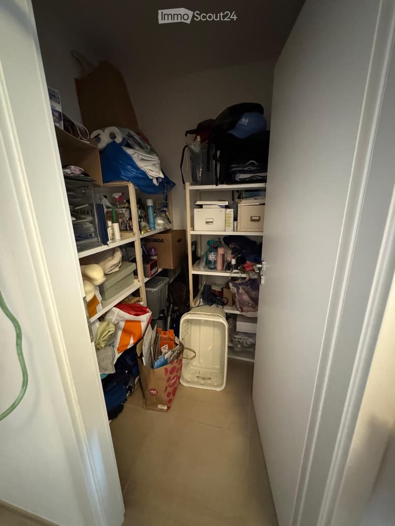 A narrow hallway with shelves on both sides filled with various items including boxes, bags, and storage containers. The floor is tiled and the walls are painted white.