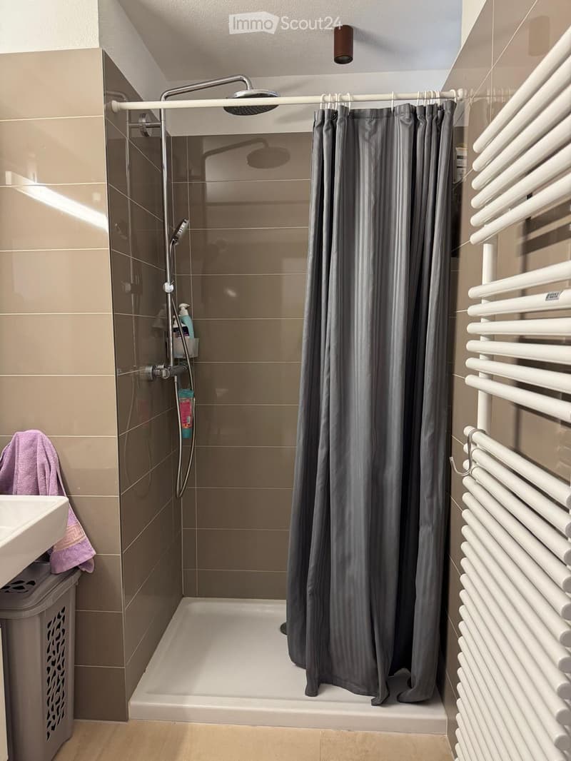 Modern bathroom with a walk-in shower, grey shower curtain, white radiator, sink, and a purple towel draped over the sink.
