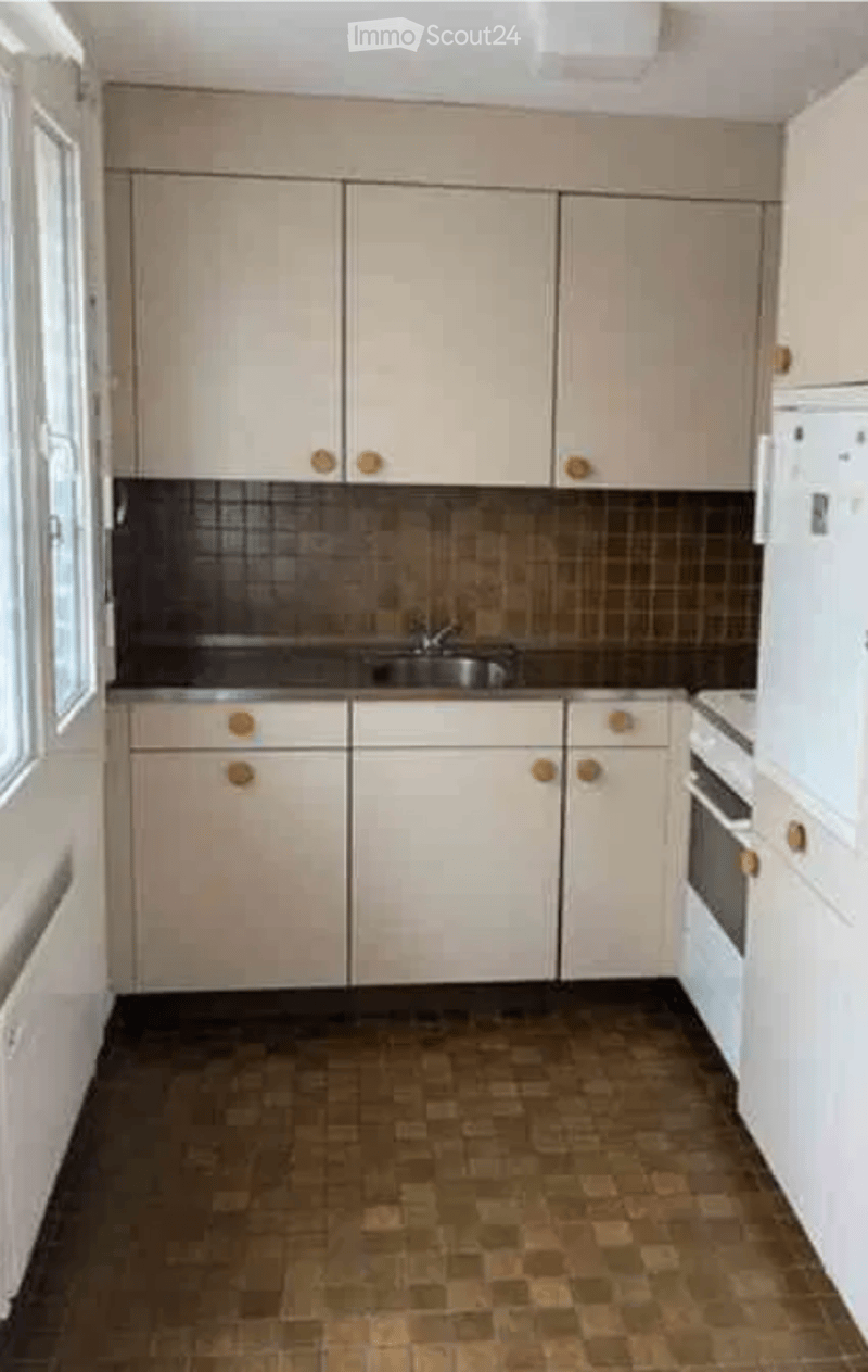 A clean, unfurnished kitchen with light-colored cabinets, a sink, and a refrigerator. The floor is tiled, and the backsplash features a dark, textured pattern.
