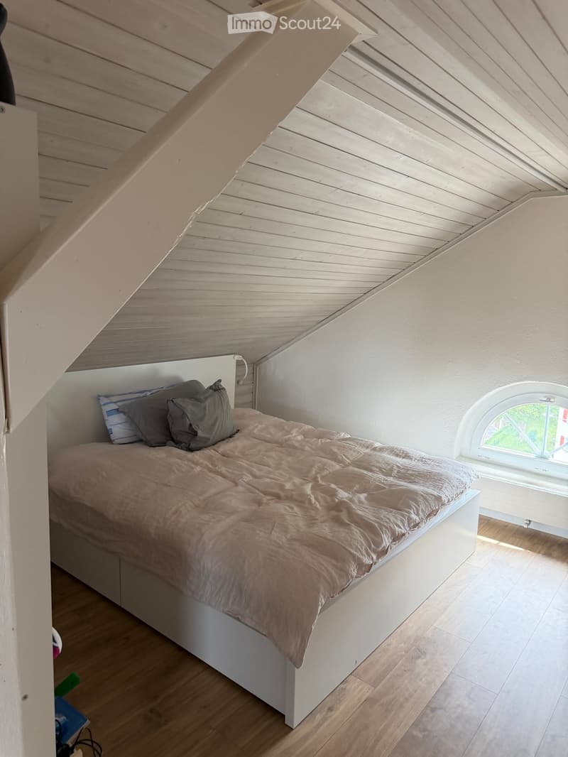 Cozy attic bedroom with wooden slanted ceilings, a white bed with a beige blanket and pillows, and a round window letting in natural light.
