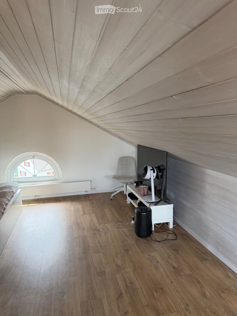 A modern, furnished room with wooden flooring, a slanted wooden ceiling, a bed, a chair, a small table with a fan and screen, and a window with a view of the outside.