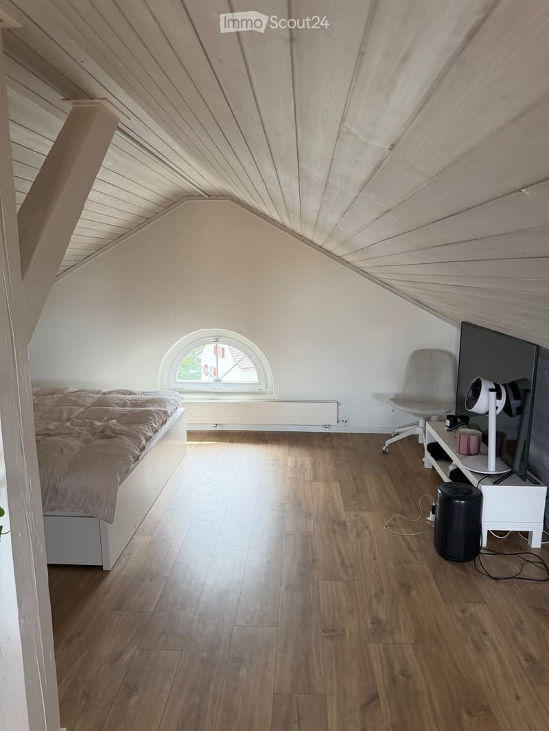 Cozy attic bedroom with wooden floors, slanted wooden ceiling, arched window, bed with beige bedding, grey chair, fan, and small table with screen.