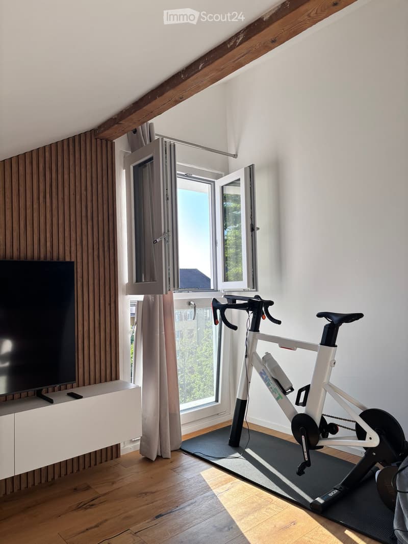 A modern, well-lit room with wooden flooring and exposed wooden beams. Features an exercise bike positioned near a large window with open blinds, a flat-screen TV on a white cabinet, and a curtain partially drawn.