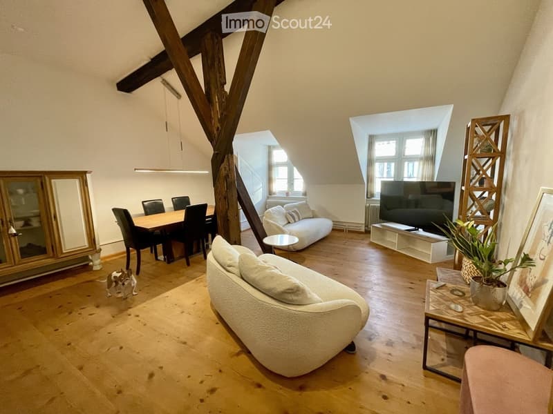 Spacious living and dining area with wooden floors, exposed wooden beams, large windows, and modern furniture including a sofa, dining table, and chairs.